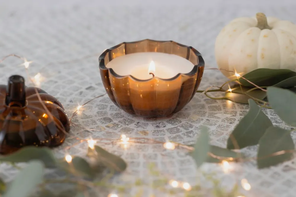 Amber pumpkin-shaped candle beside a small white pumpkin and autumn greenery.