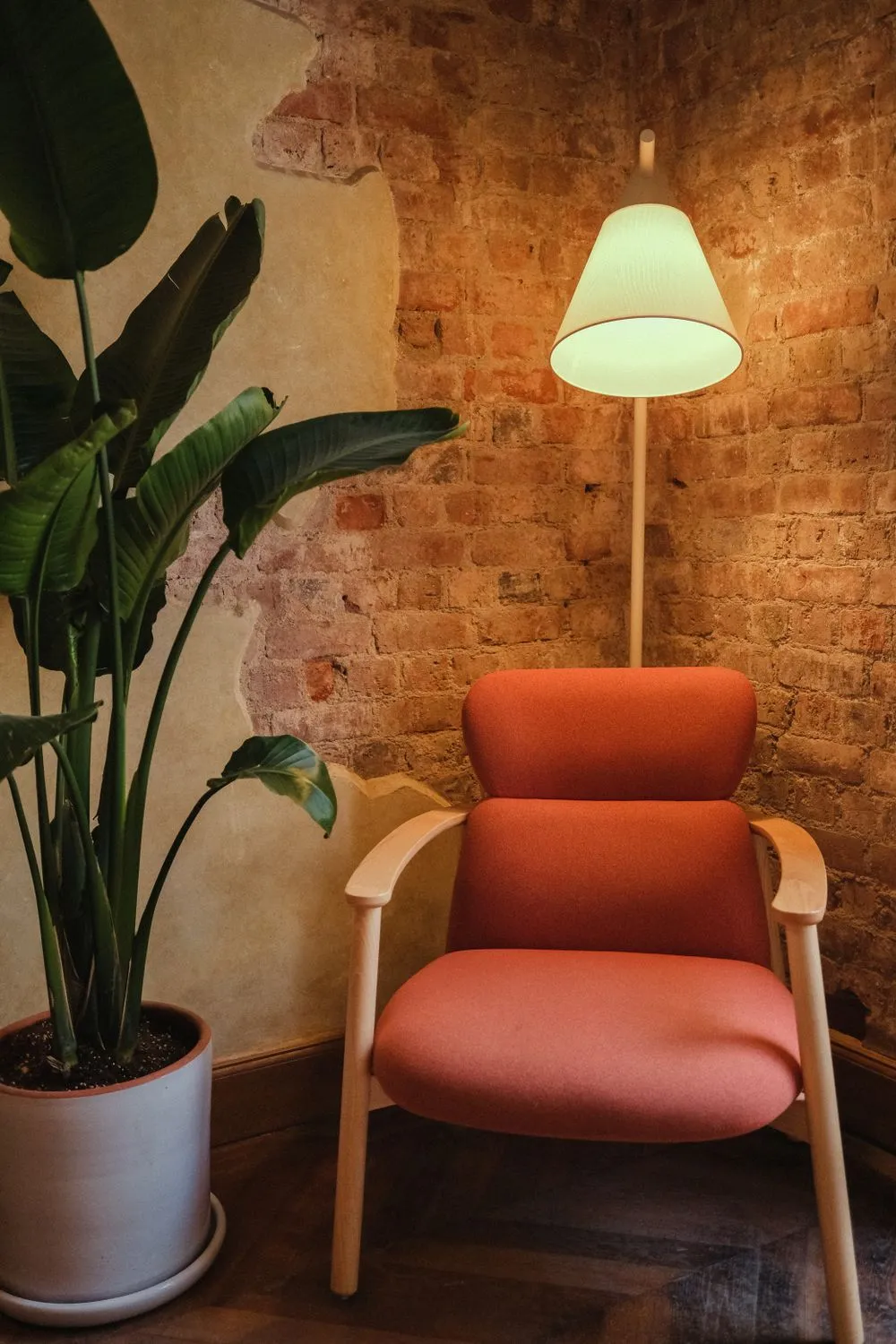 Rust-colored armchair beneath a floor lamp beside a leafy plant and exposed brick wall.