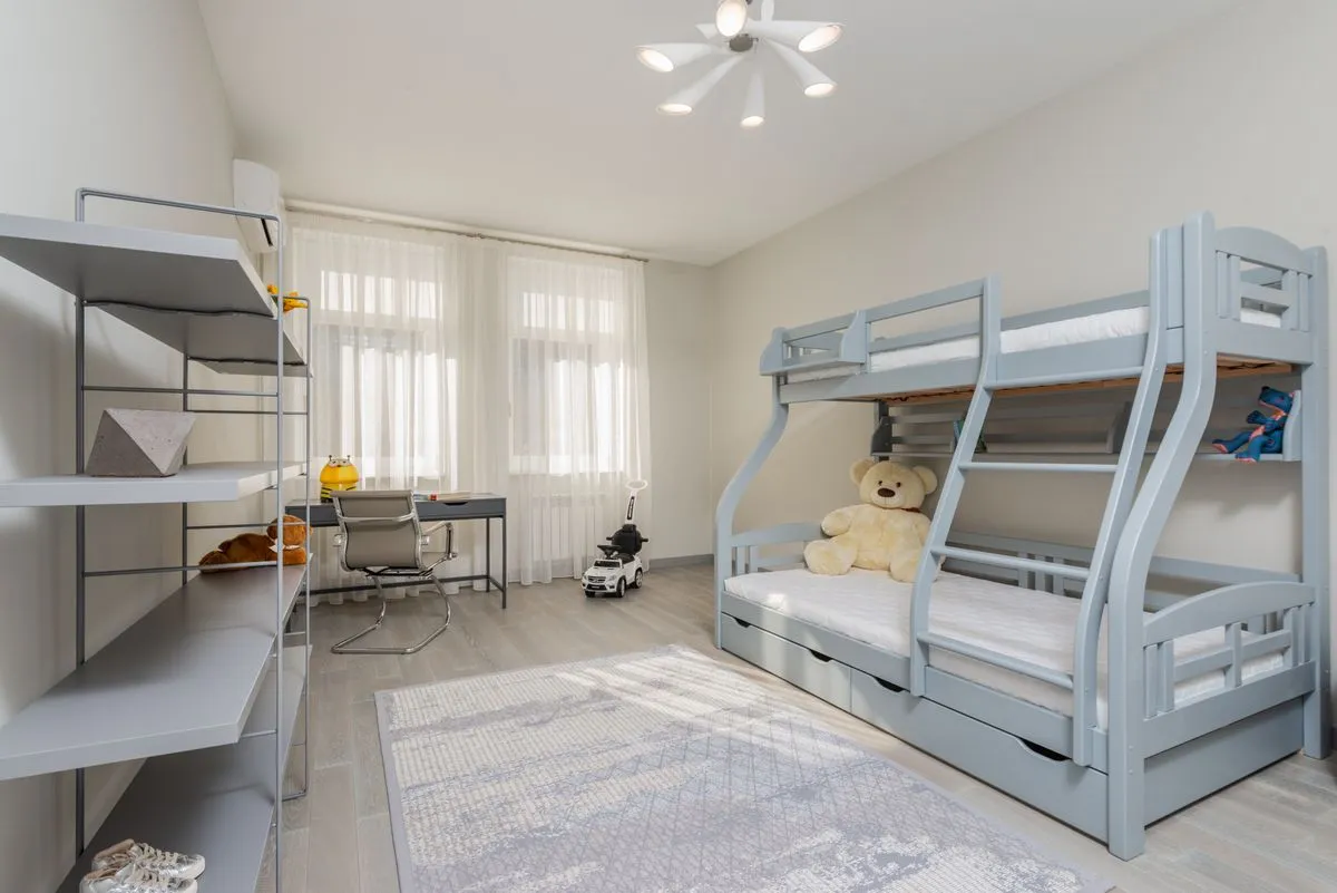 Pale blue bunk bed with under-bed drawers, open shelves, and a desk beside a bright window.
