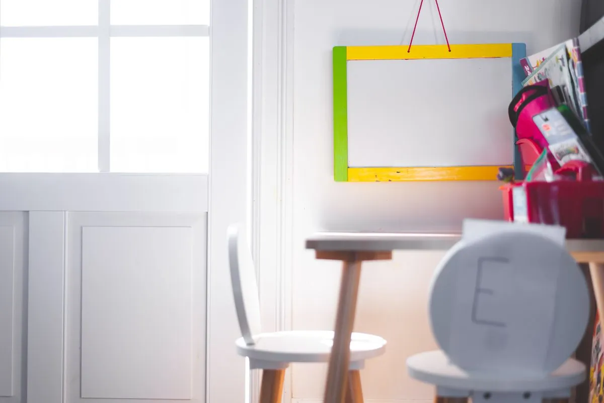Small wooden desk with white chairs and a colorful drawing board beside a window.